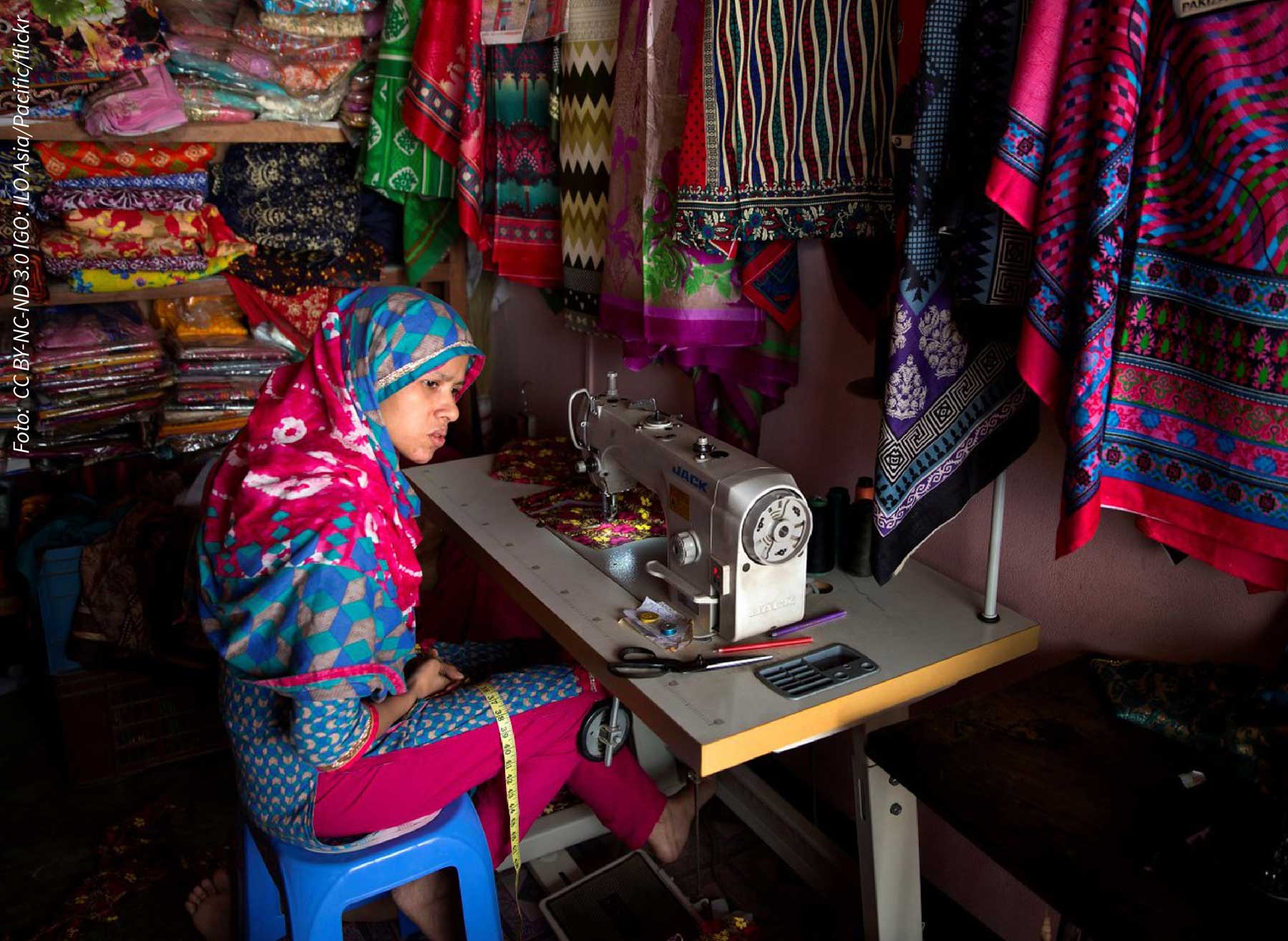 Eine Näherin sitzt vor einer Nähmaschine in einem mit buntem Tuch angefüllten kleinen Raum. 