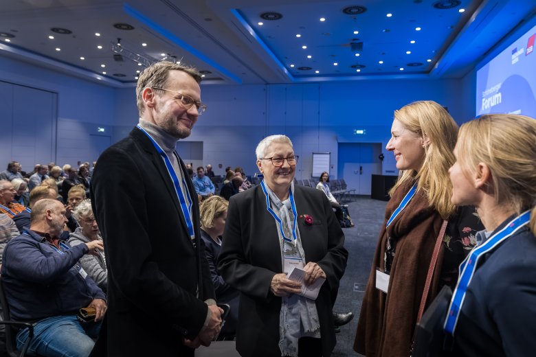 Eine Tagungssituation. 4 lächelnde Menschen unterhalten sich im Vordergrund. Im Hintergrund Sitzreihen mit Menschen. 