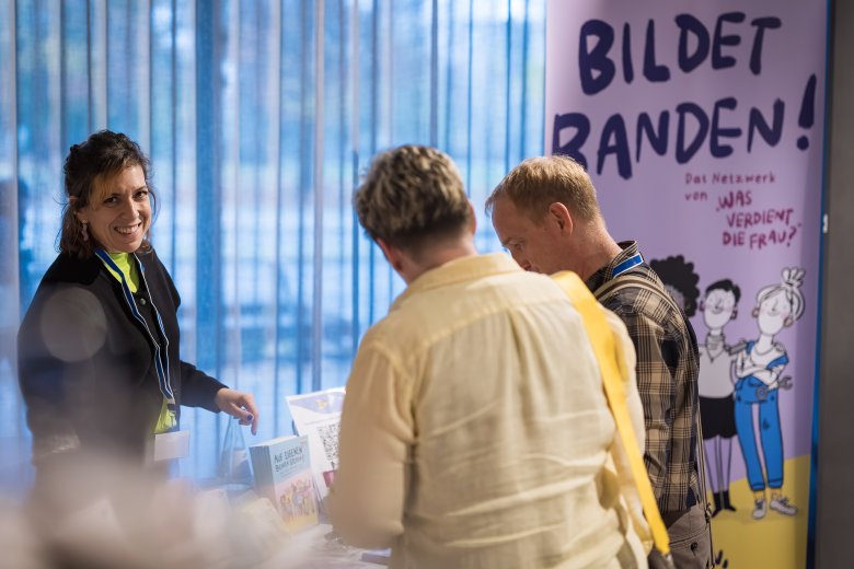 Ein Infostand mit einer lachenden Frau hinter dem Infotisch und 2 interessierten Menschen davor. Im Hintergrund in Banner auf dem steht: Was verdient die Frau?