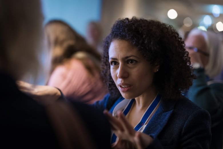 Das Gesicht einer Frau, die diskutiert. Sie trägt ein Namensschild-Band um den Hals. Im Hintergrund Menschen, die unscharf zu sehen sind..