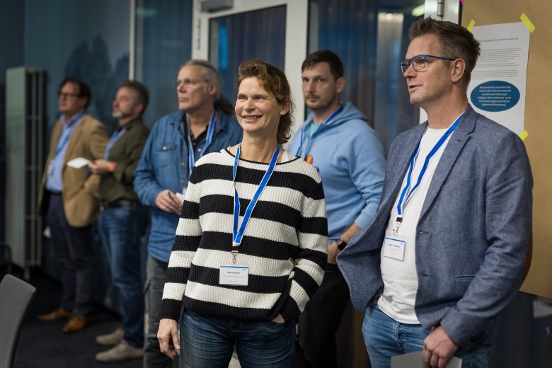 Eine Gruppe von 5 Menschen in einem Raum. Sie schauen in eine Richtung und tragen Namensschilder um den Hals. Im Hintergrund sind Infowände zu sehen.