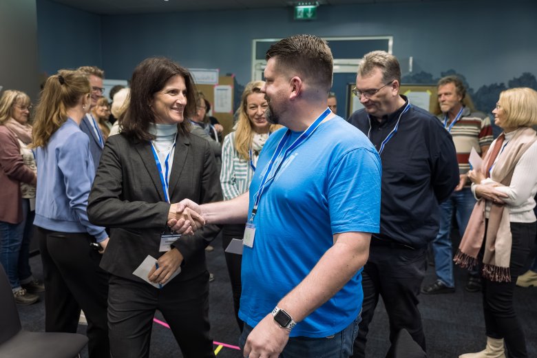 Eine Frau und ein Mann stehen in einem Raum vor einer Gruppe von Menschen. Sie schauen sich in die Augen und schütteln sich die Hand. Im Hintergrund sieht man Infowände.