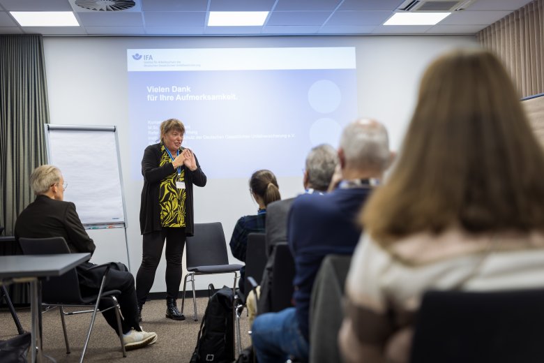 Ein Tagungsraum mit einer Rednerin vor einer Leinwand. Das Publikum auf Stuhlreihen sieht man von hinten.