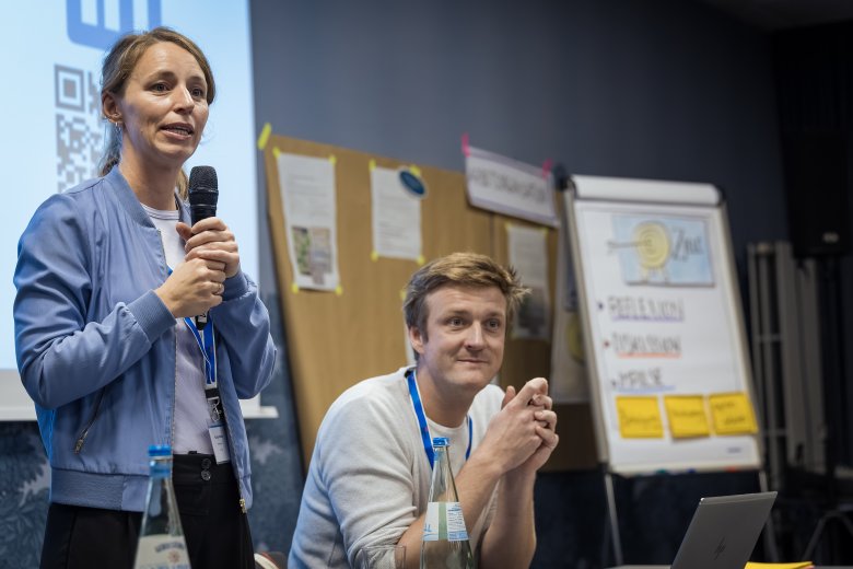Eine stehende junge Frau mit einem Mikro in der Hand, die einen Vortrag hält. Neben ihr sitzt ein junger Mann. Im Hintergrund Infowände und ein Flipchart.
