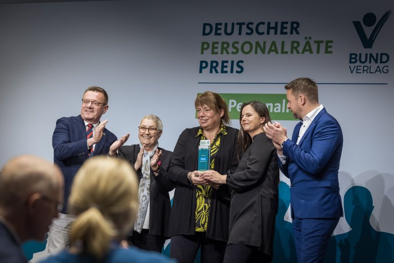 5 lachende und applaudierende Menschen vor einer Leinwand mit dem Schriftzug "Deutscher Personalrätepreis" und einem Logo des Bund-Verlags.
