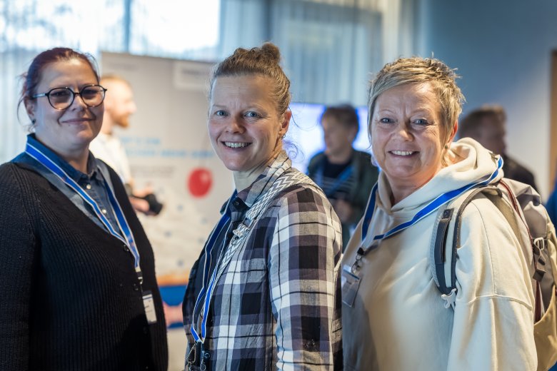 Drei lachende Frauen, die in die Kamera schauen und Namensschilder um den Hals tragen. Im Hintergrund Infotafeln.