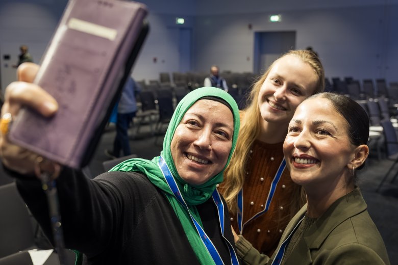Drei lachende Frauen von vorne, die ein Selfie machen. Im Hintergrund ein Raum mit leeren Stuhlreihen.