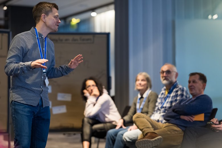 Ein junger Mann, der einen Vortrag hält. Er trägt ein Namensschild um den Hals. Unscharf im Hintergrund eine Infowand mit Text. und eine Gruppe von 4 Menschen die zuhören.