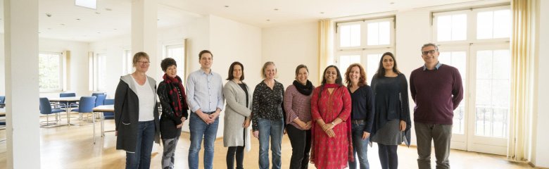 10 Personen, 8 weiblich, 2 männlich, überwiegend leger gekleidet, eine Person in asiatischer Kleidung, stehen im Halbkreis in einem lichtdurchfluteten Raum, der einem Seminarraum vorgelagert ist. Alle blicken freundlich lächelnd in Richtung des/der Bildbetrachters/Bildbetrachterin. 