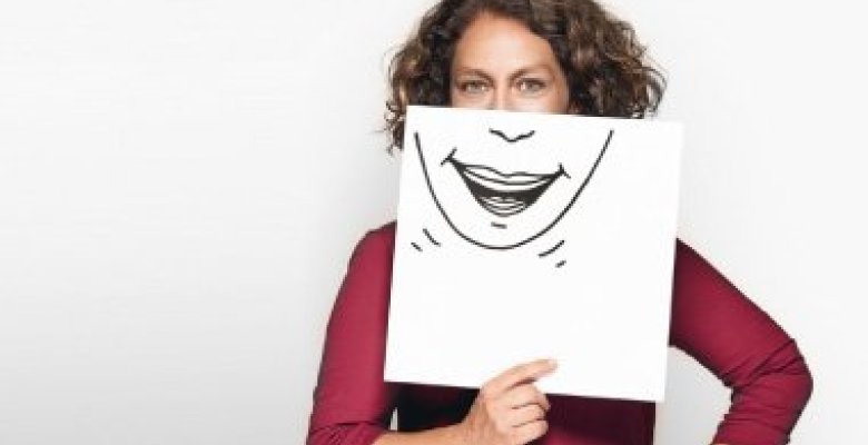 Eine Frau ist in dem Bild zu sehen. Sie hält ein Blatt vor ihr Gesicht. Auf diesem Gesicht ist ein lachender Mund gemalt