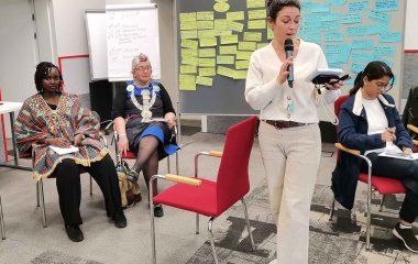 Vier Frauen in einem Seminarraum im Tagungszentrum Hattingen. Drei sitzend, die Vierte, im Vordergrund des Bildes, ist offensichtlich in Bewegung, während sie in ein Mikro spricht. Im Hintergrund: 2 Stellwände, bestückt mit beschrifteten rechteckigen Moderationskarten, die einen in Gelb, die anderen in Blau.